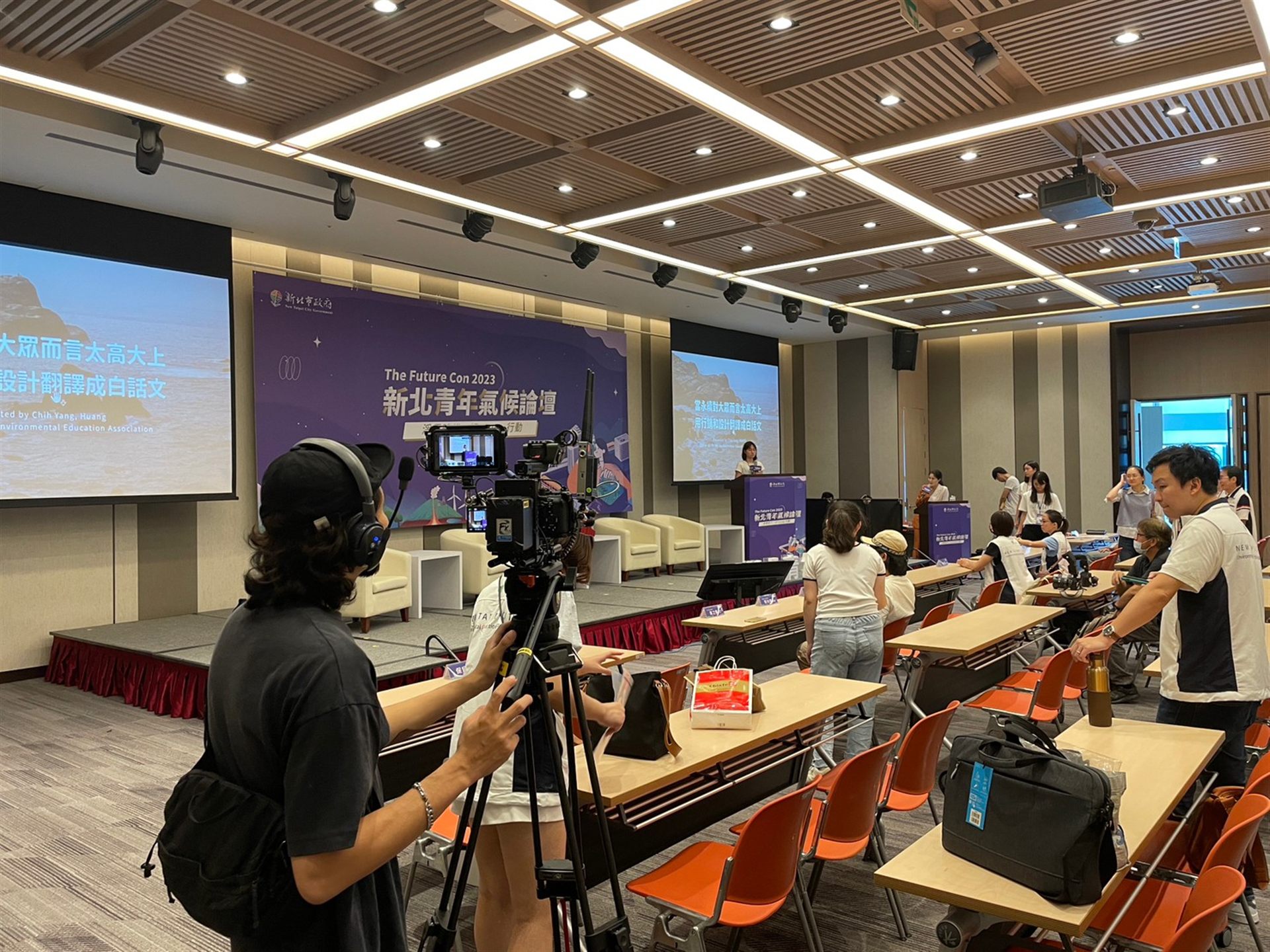 新北青年氣候論壇現場直播側拍:攝影師手持攝影機拍攝舞台講者與雙螢幕活動視覺背板